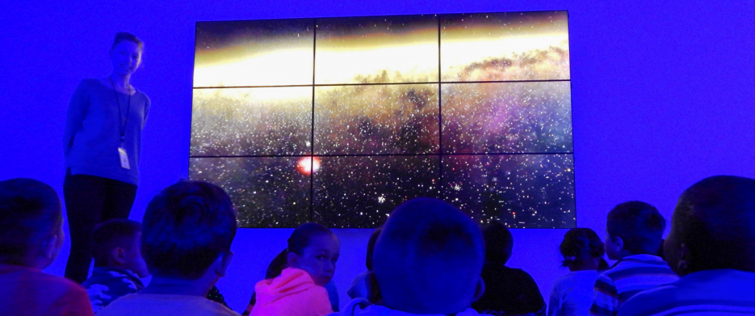 Students in a dark room look up at nine screens arranged in a grid, displaying what could be the universe or perhaps dust under a microscope. One of the girls, in a pink shirt, looks away from the screen and at the viewer.