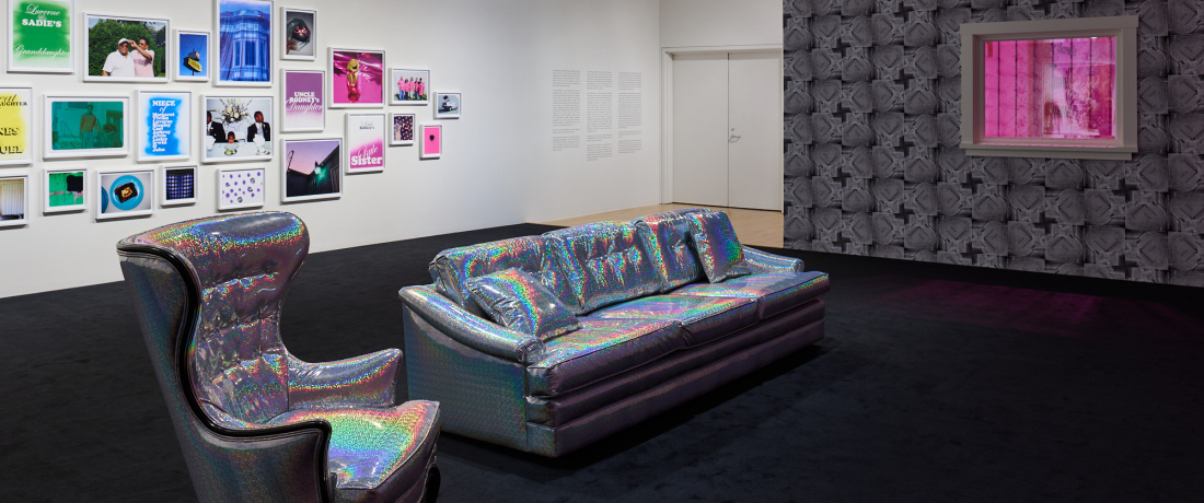 A shiny silver couch and armchair face a wall that is not pictured. A window with bars on it is cut into a wall that has a geometric black and grey wallpaper. Outside the window is a pink glow. Another wall has dozens of colorful framed photos. 