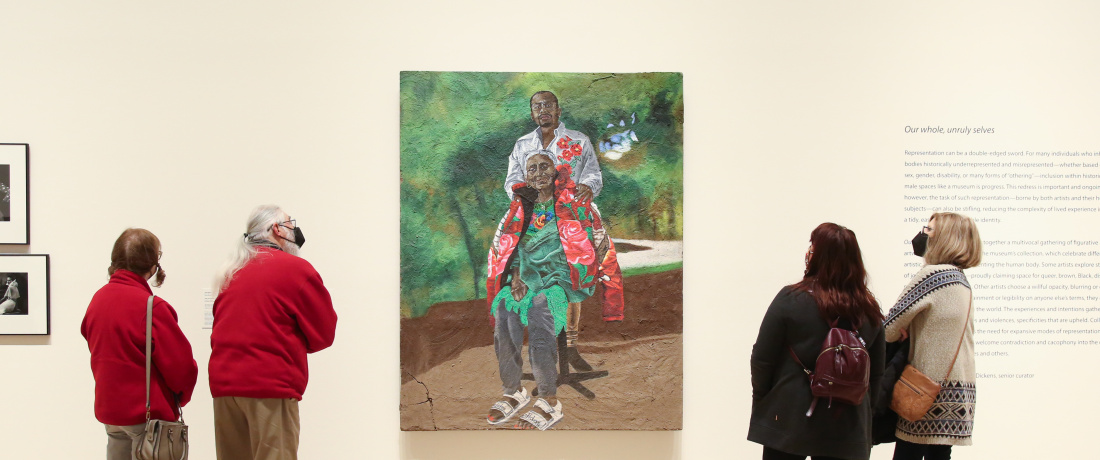 Two couples stand on either side of an oversized painting. The portrait is of an older woman with grey and white hair and brown skin who is wearing a floral jacket and adidas sandals. Behind her with his hand on her shoulder, is a young man with dark skin wearing a white shirt. 
