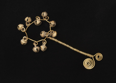 Against a black background is a baby brass rattler. It looks to be constructed from a single piece of brass wire, which is looped around itself to make a handle. Extending from it is a circle made of small circles that each have a bell looped through them. 