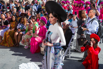 A slender Vietnamese woman wearing a custom ao dai dress with a large round hat in front of a large packed crowd.