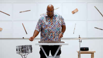 Man in a colorful shirt uses drum sticks to hit percussion instruments, including a snare drum and jail bars that are placed on a keyboard stand.