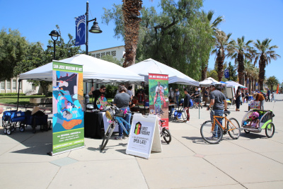 an outdoor festival event with a booth of two large white canopies and stand-up banners of artworks with people in bikes talking to a museum staff.
