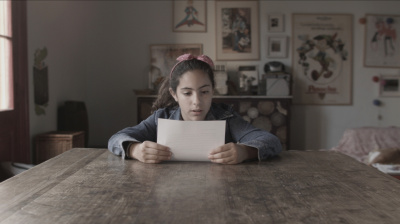 A young girl with a blue denim jacket and pink headband sits at a large table and reads from a sheet of paper. 