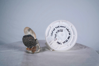 On a table lay two objects: one a cement piece with a coin and round rock sticking out of it and the other the plastic lid of a container with words handwritten on it.