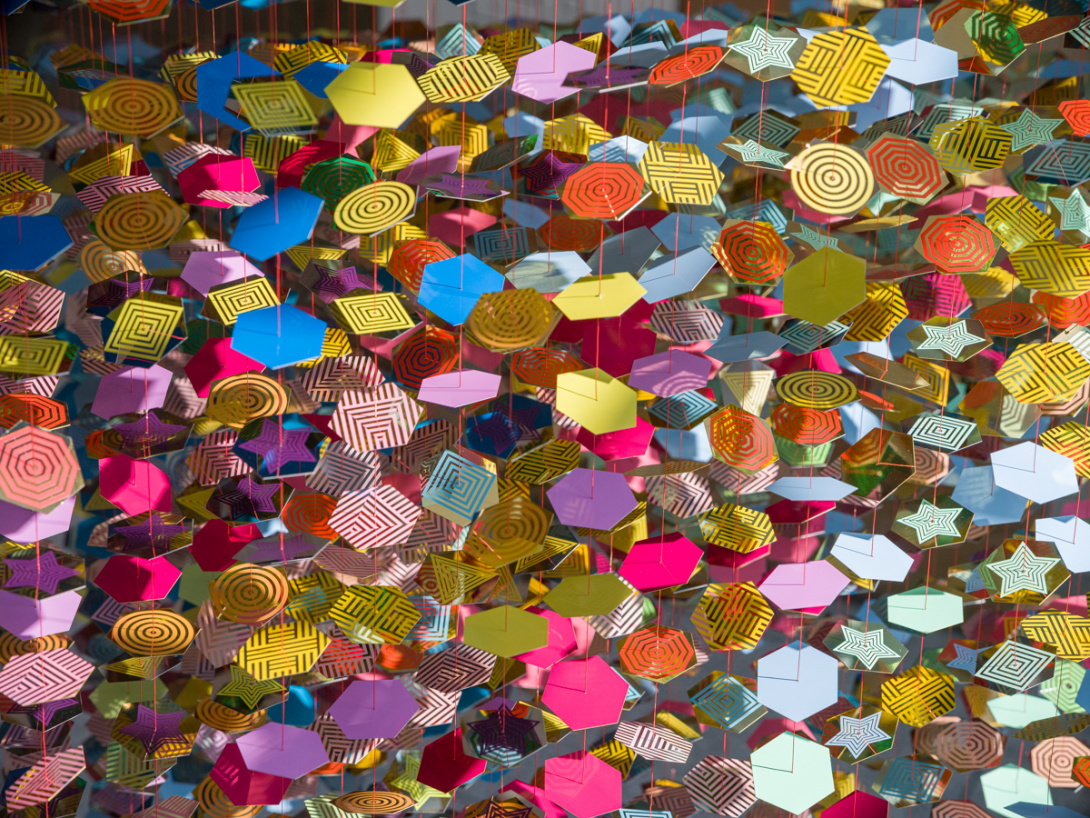 A close-up view of hexagonal disks which they are aligned with neon orange cables and suspended from above. Reflective and shimmering disks are gradated colors of pink, yellow, orange, blue, and purple.