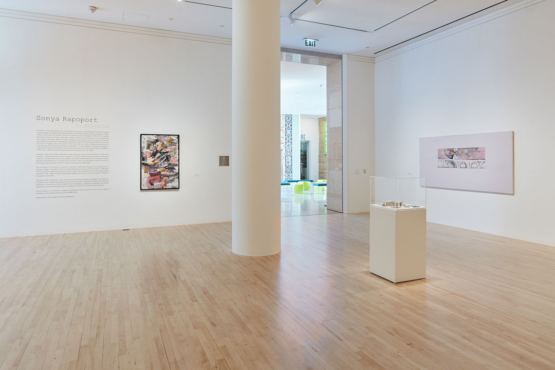 A gallery space with cream walls and birch wood flooring. There are texts, an abstract painting, and a cylinder building column along with a painting. In the background is an open entrance. In the foreground is an artwork on a pedestal with a transparent case.