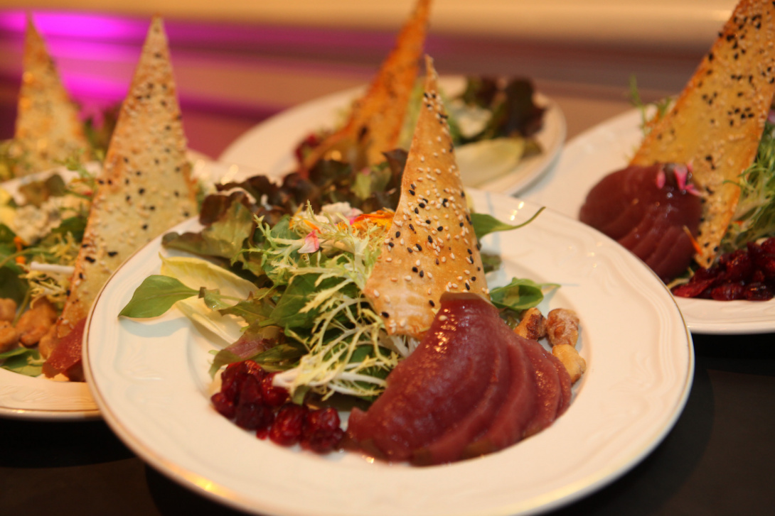 Small plates hold triangular flatbreads which sit atop mixed greens with rocket lettuce, with hazelnuts and sliced beets on the side. The plate in front is on sharp focus, while those in the back are blurred. Magenta light shines in the background. 