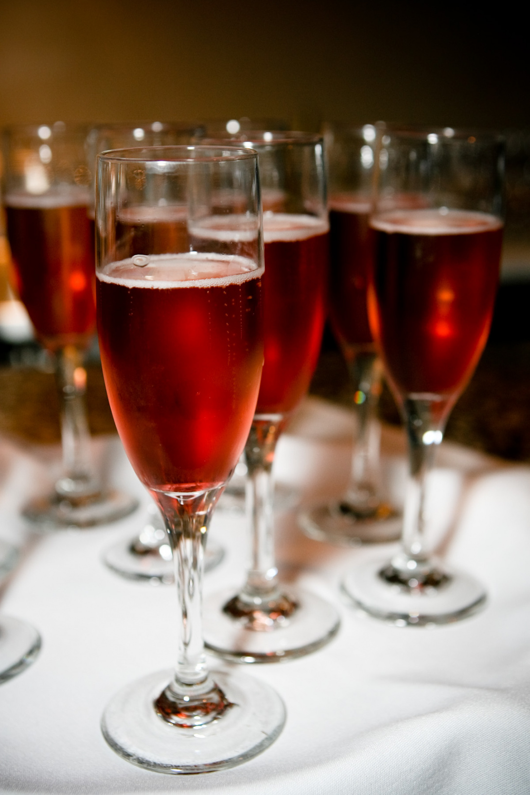 A series of glass champagne flutes with a cranberry-colored fizzy liquid bubbling away. One glass in the foreground is in sharp focus, 4–5 others stand behind it. The glasses sit on a tablecloth, the table’s edge can just be seen behind the glasses. 