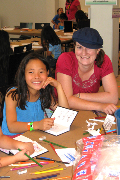 A woman with brown hair in pig tales wearing a blue hat and a pink shirt is smiling next to a smiling child wearing a blue tank top writing in a book. They are in a room filled with tables and art supplies. Other children and adults are in the distance. 