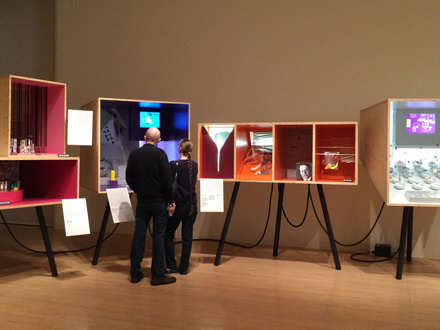 A room with a man and woman looking at displays. From left to right is a portrait box stacked on top of a horizontal box, second is a large square box, third is a large horizontal rectangular box sectioned into 4 spaces, the fourth is a large square box.