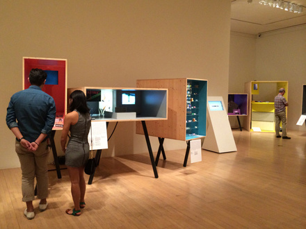 A gallery with 3 people looking at displays. They are of various sizes and orientations: with red lining inside, with a small projection, an upright rectangular box, a white stand with a small screen, a purple box, and a divided yellow box.