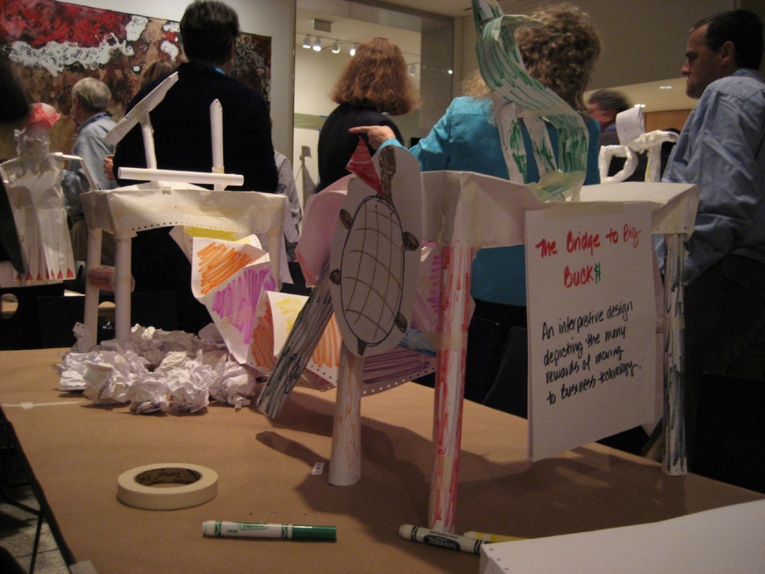 A group of adults stands facing away from the photo. On the table there are crafts made out of paper in the shape of animals, bridges, and cliffs. There is also tape, and Crayola markers.