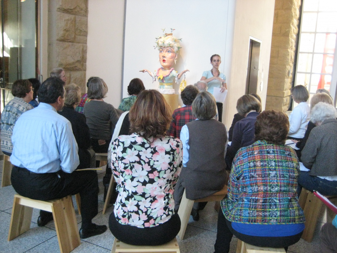 A docent stands in front of a crowd seated on wooden stools leads a discussion. Next to her is a sculpture of a Victorian woman directly to her right.