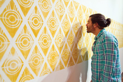 A bright yellow horizontal band comprised of diamonds with white concentric circles inside of them. A visitor looks closely at the work. The work is quite close on the left side and fades into the distance on the right behind the visitor. 