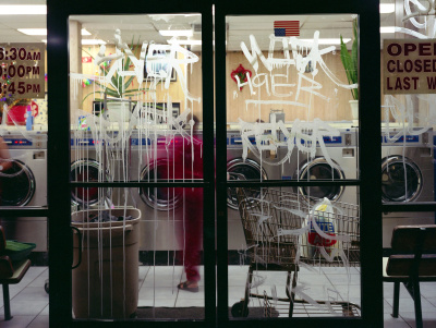 a photo of a coin wash facility from the outside with the glass covered in graffiti and a woman inside wearing a sweatsuit and cart with a bag of clothes